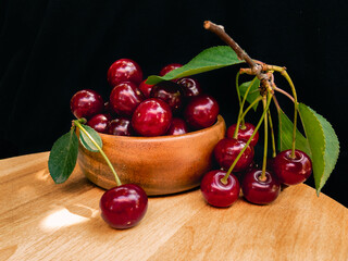 Juicy ripe red cherries with green leaves in a wooden bowl on a wooden surface on a black background.