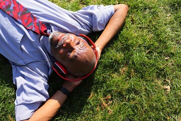 African American man in blue shirt and red tie lays in the grass listening to music through red...