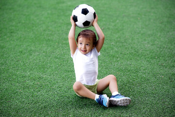 Toddler boy in sports uniform sitting on green footall field holding soccer ball above head. Little child playing with ball on grass. Active childhood and outdoor games for children concept