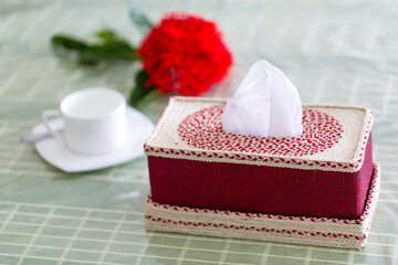 Deep red Handmade fiber Napkin box on the dining table with a red flower and a white tea-cup. Tissue paper box on restaurant.