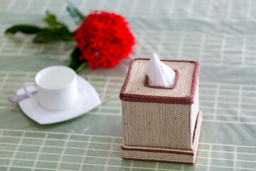 Raw fiber color handmade Napkin box on the dining table with a red flower and a white tea-cup. Tissue paper box on restaurant.