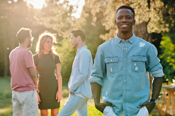 Happy young African man in denim shirt standing in park against friends