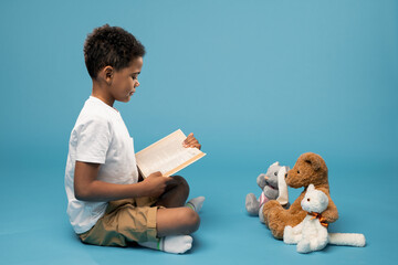 Serious and diligent African schoolboy reading book of tales to his toys