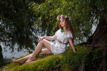 Naklejka na ściany i meble Beautiful girl in white dress and flowers wreath on green willow background. 7th July, traditional slavic holiday with fortune-telling and divination rituals in Ukraine.