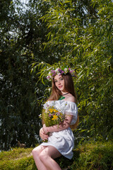 Beautiful girl in white dress and wreath holding field flowers bouquet on green willow background. 7th July, traditional slavic holiday with fortune-telling and divination rituals in Ukraine.