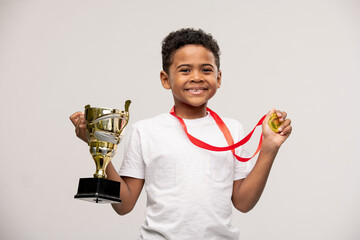 Joyful cute mixed-race little boy holding medal and golden cup in hands