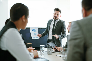 Confident broker showing paper with financial diagram to colleagues at meeting