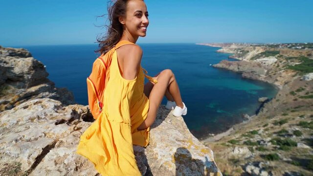 taned traveler woman sitting on cliff top in front of amazing seascape, bright yellow dress and hair blowing in the wind. Freedom, travel and vacation concept.