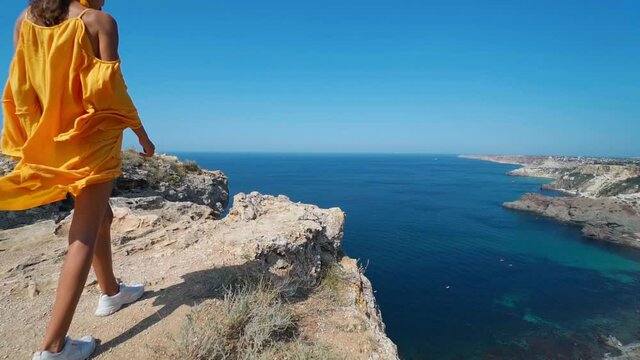 Full length epic slow motion of taned traveler woman walking on top rock cliff in front of amazing seascape, bright yellow dress and hair blowing in the wind.
