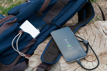 Modern smartphone are charging from power bank lies next to the backpack on a wooden stump. Modern...