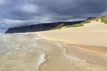 Polihale State Park in Kauai, Hawaii. The view on the empty white sandy beach and clear waters of Pacific ocean. Ha‘Ele‘Ele Valley and dark sky in the background.