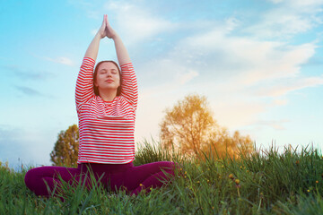 A girl in a striped t-shirt practices yoga at sunset. Blue sky and green grass. The woman is sitting on the ground with her hands up, palms folded and eyes closed.