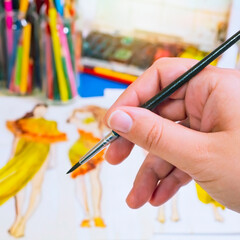 The process of drawing and creativity. The artist holds a thin brush in his hand against the background of his sketch in watercolors. The picture is square.