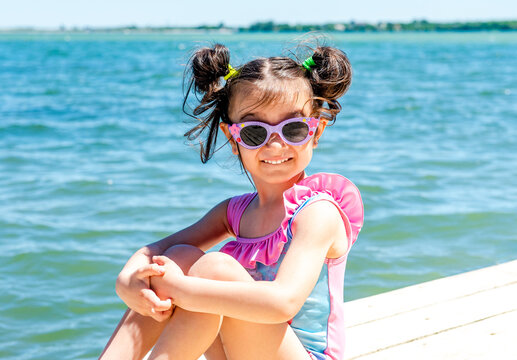 Little cheerful girl in a swimsuit and sunglasses sits on a wooden pier on the lake against the background of blue water. Baby girl smiling while sitting on a pier