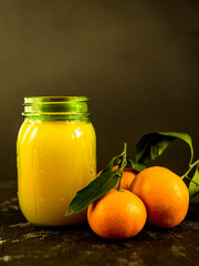 Fresh tangerine juice in a glass jar on a black background. Whole tangerines with green leaves lie nearby. Healthy eating concept