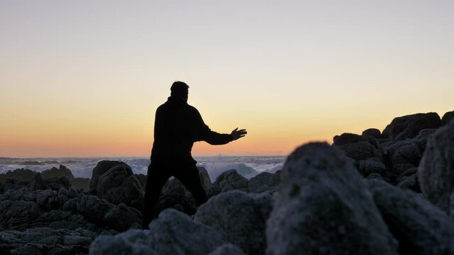 Men warrior monk practicing silhouette tai chi karate kung Fu on the rocky stones horizon at sunset or sunrise. Art of self-defense. Silhouette on a background of dramatic epic waves at pacific coast
