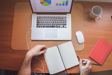 Office flat lay with man writing on a notebook and working with a laptop. The screen on the laptop has reports and graphics. Work from home concept. Business person and freelance.