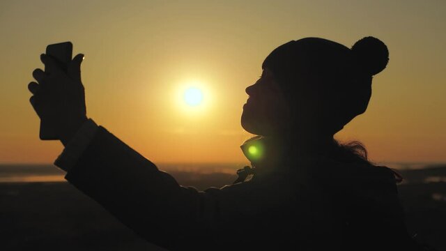 silhouette of a woman in sunshine, selfie, taking pictures at sunset, sunrise. Free Young girl tourist blogger records a selfie video on top of mountains using a smartphone with beautiful landscape.