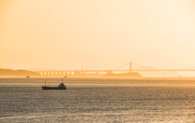 Cargo ship cruising the shores of Naoshima Island, Japan, at the misty sunset with the Great Seto Bridge seen in the background.