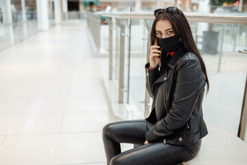 Girl with medical black mask and mobile phone in a shopping center. Coronavirus pandemic. A woman with a mask is standing in a shopping center. A girl in a protective mask is shopping at the mall