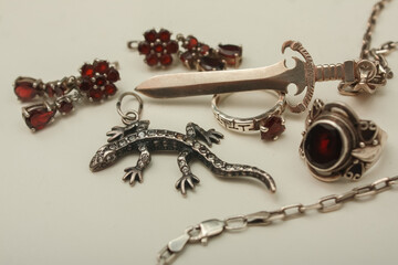 Silver pendant in the shape of a sword on an anchor chain, silver set of earrings and rings with natural red garnet stone, pendant in the shape of a lizard with transparent zircon on a white backgroun