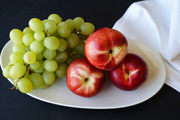 Fresh nectarines and green grapes on white plate, linen napkin, black background, elegant summer fruit still life.