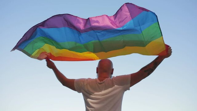 Homosexual bold man with rainbow gay flag while parade on background of blue sky. LGBTQI, Pride Event, LBGT Pride Month, Gay Pride Symbol