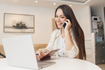 Young woman with baby speaks by phone and working with laptop on kitchen. Business Quarantine Concept
