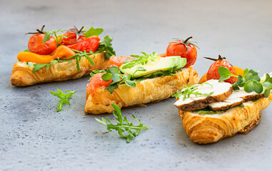 Variety of croissant sandwiches with grilled pepper, tomatoes, smoked salmon, turkey, avocado and arugula served with micro green on grey concrete background. Close up.