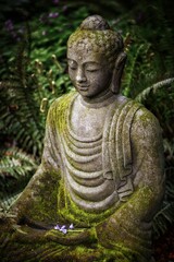 Vertical shot of a Buddha statue with moss on top and greenery on the background