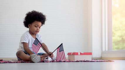 Black African boy sitting next to gift box, on the floor holding USA flag. 4th of July concept.