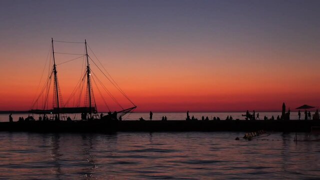 Peaceful sunset with a high contrast view of a two-masted boat by the pier, on a crowded and lively day.