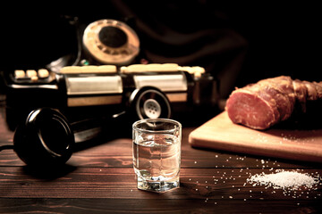 A vodka glass on an old table with an old black phone meat on the board with salt with shallow depth of field and blur.