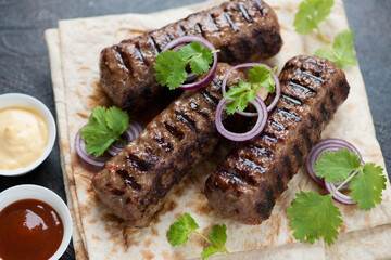 Grilled kebabs made of marbled beef meat with lavash, fresh cilantro, red onion and dips, close-up, selective focus