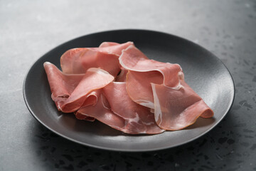 Thin italian prosciutto slices on black plate on conctere background