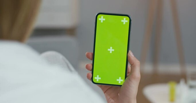 View over shoulder of woman on black vertical smartphone with green screen and tracking motion. Girl sitting at home wrapped in blanket and watching video on cellphone. Chroma key. Indoor.