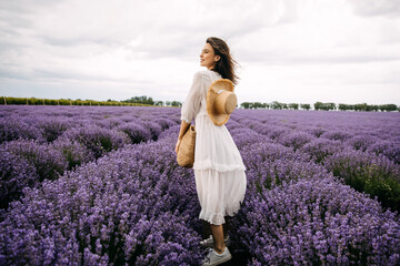 Young romantic woman standing in lavender field, wearing a bohemian white maxi dress and a straw...