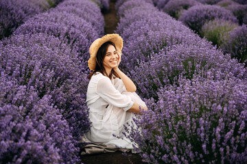 Young brunette woman sitting between rows of lavender bushes, smiling, wearing a bohemian white...