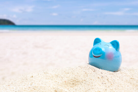 Summer happy piggy bank on sand beach over blurred tropical blue sea background, image for saving vacation concept.