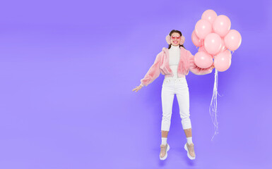 Horizontal banner of young happy girl, wearing winter clothes and ear warmers and jumping with pink helium birthday balloons, isolated on blue background with copy space