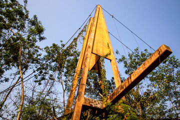Part of yellow metal suspension bridge being abandoned in the wild. Metal structure of a bridge against green tree background.
