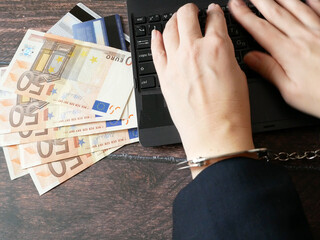 unknown person hands in business suit with handcuffs and money near on table works with laptop near...