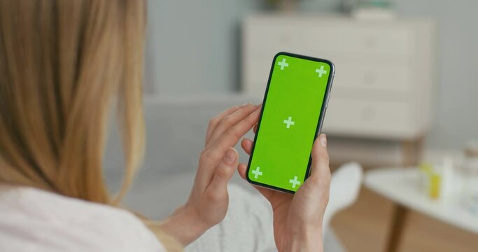 View over shoulder of woman on black vertical smartphone with green screen and tracking motion. Girl lying on couch at home and tapping or scrolling news page on mobile phone. Close up.