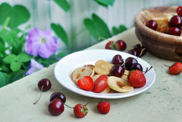 Homemade Mini pancakes and fresh strawberries, cherries on an old wooden table. Garden flowers in the background.