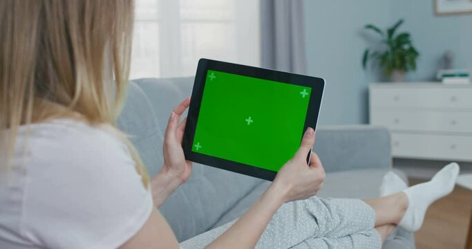 View over shoulder of woman on black horizontal tablet computer with green screen and tracking motion. Girl lying on couch at home and tapping or scrolling news page on gadget with chroma key Close up