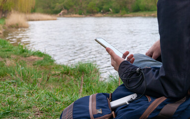 Man uses smartphone while charging from the power bank on the river's bank. Modern technology concept.