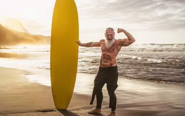 Happy fit senior having fun surfing at sunset time - Sporty bearded man training with surfboard on the beach - Elderly healthy people lifestyle and extreme sport concept