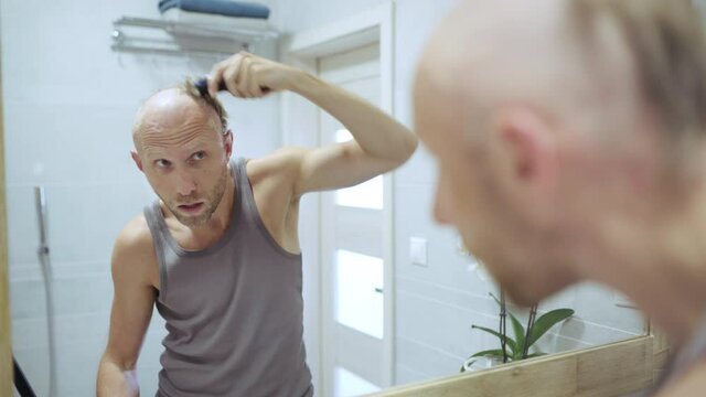 4K Man shaving his hair completely bald in front of bathroom mirror with electric razor. Hairdresser concept, self-sustainability concept, asceticism concept.