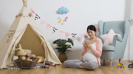 asian female shopping online with mobile phone in her child’s playroom. happy mother-to-be seated...