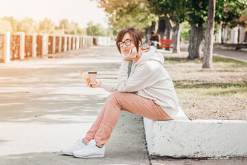 Woman in white hoodie with paper cup of coffee is sitting outside in autumn park.